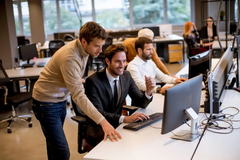 Colleagues collaborate at a desk with a computer, one man in a suit smiling as others assist him.